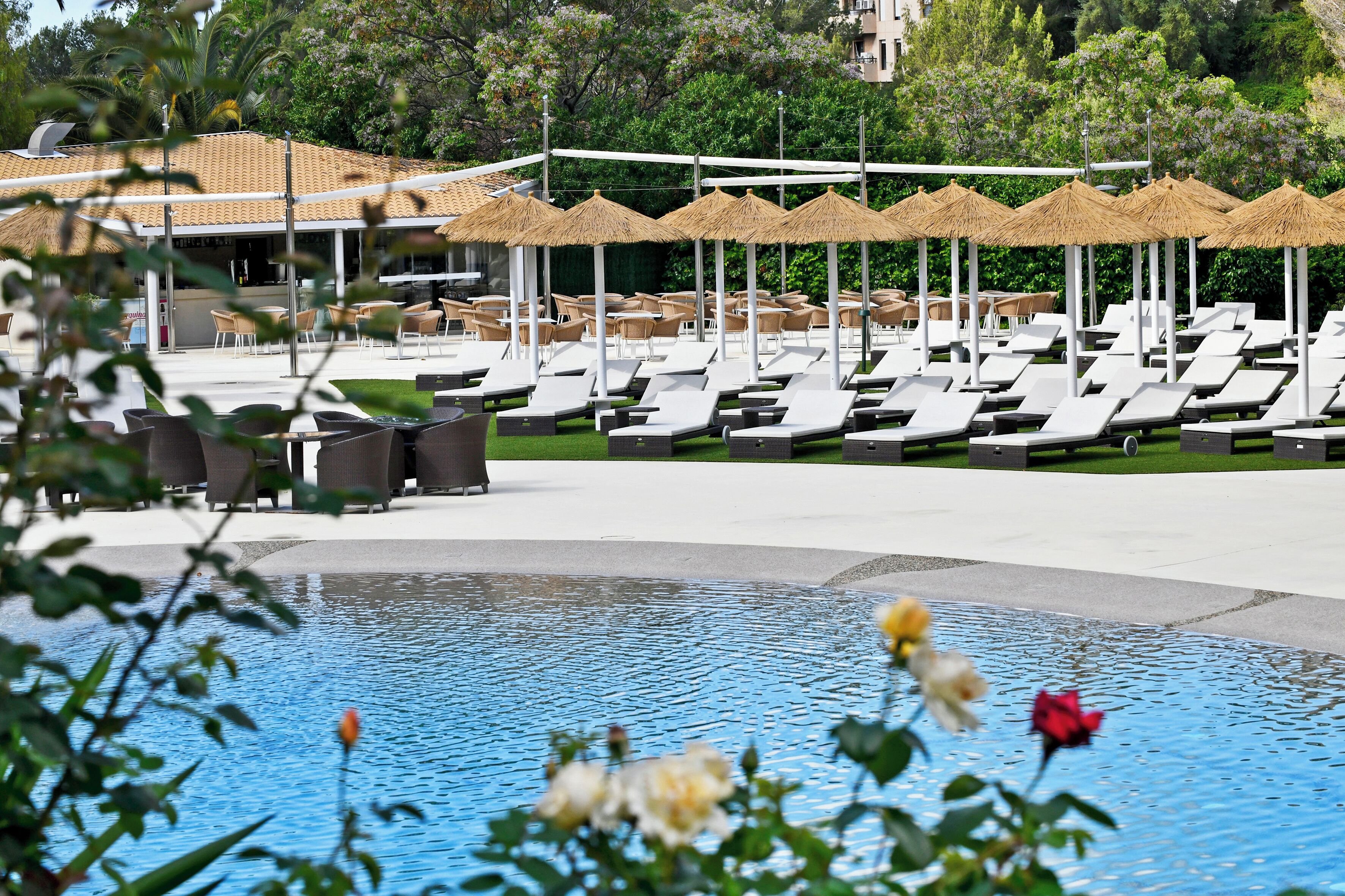 Zona de piscina con sillones de descanso y sombrillas, rodeada de vegetación y un edificio con tejado de tejas.