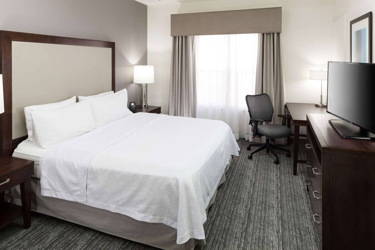 A hotel room with a large bed, a desk with a chair, a television, and a window with curtains.