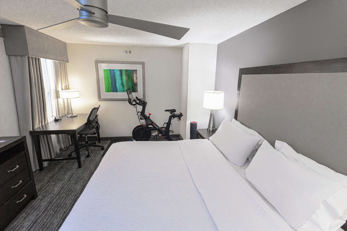 A hotel room with a large bed, a desk, a chair, a bicycle, and a ceiling fan.
