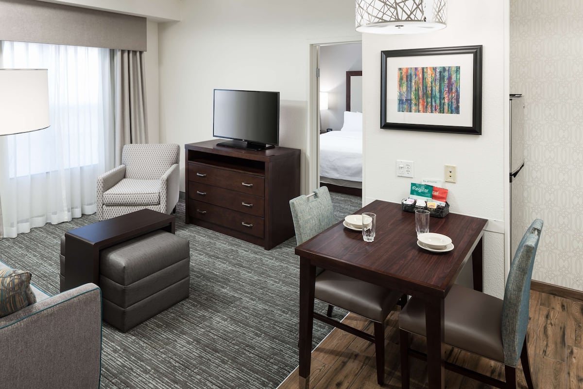A hotel room with a TV on a dresser, a dining table with chairs, a sofa, and a bed in the adjacent room.