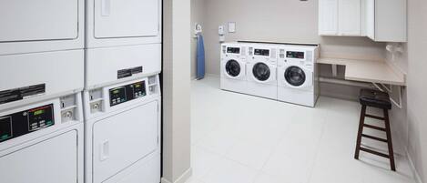 A laundry room with multiple washing machines and dryers, a blue ironing board, and a wooden stool.