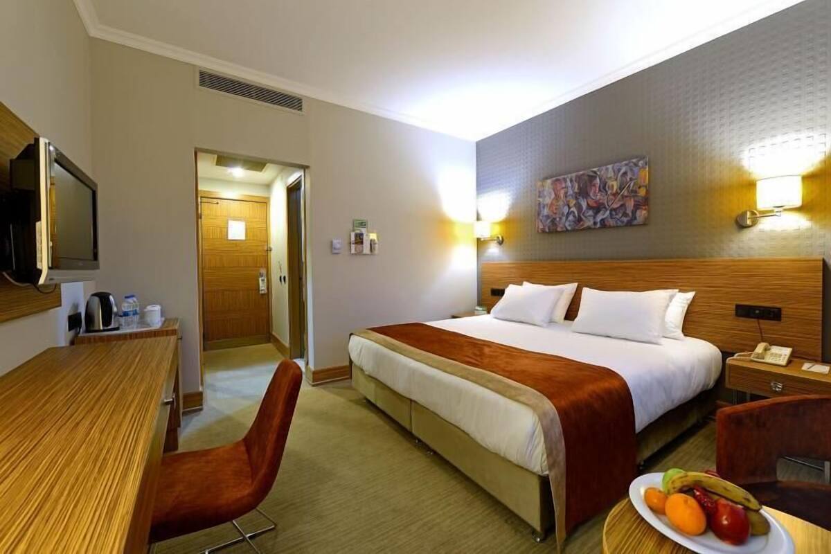 A hotel room with a bed, a desk, a chair, a bedside table, a fruit plate, and a painting on the wall.