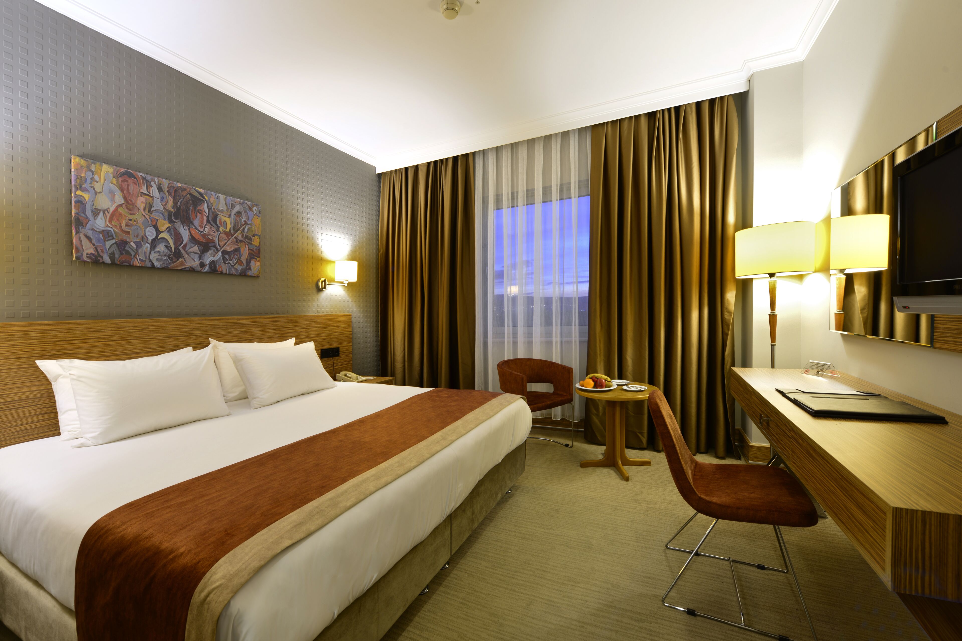 A hotel room with a large bed, a desk with a chair, a television, and a painting on the wall.