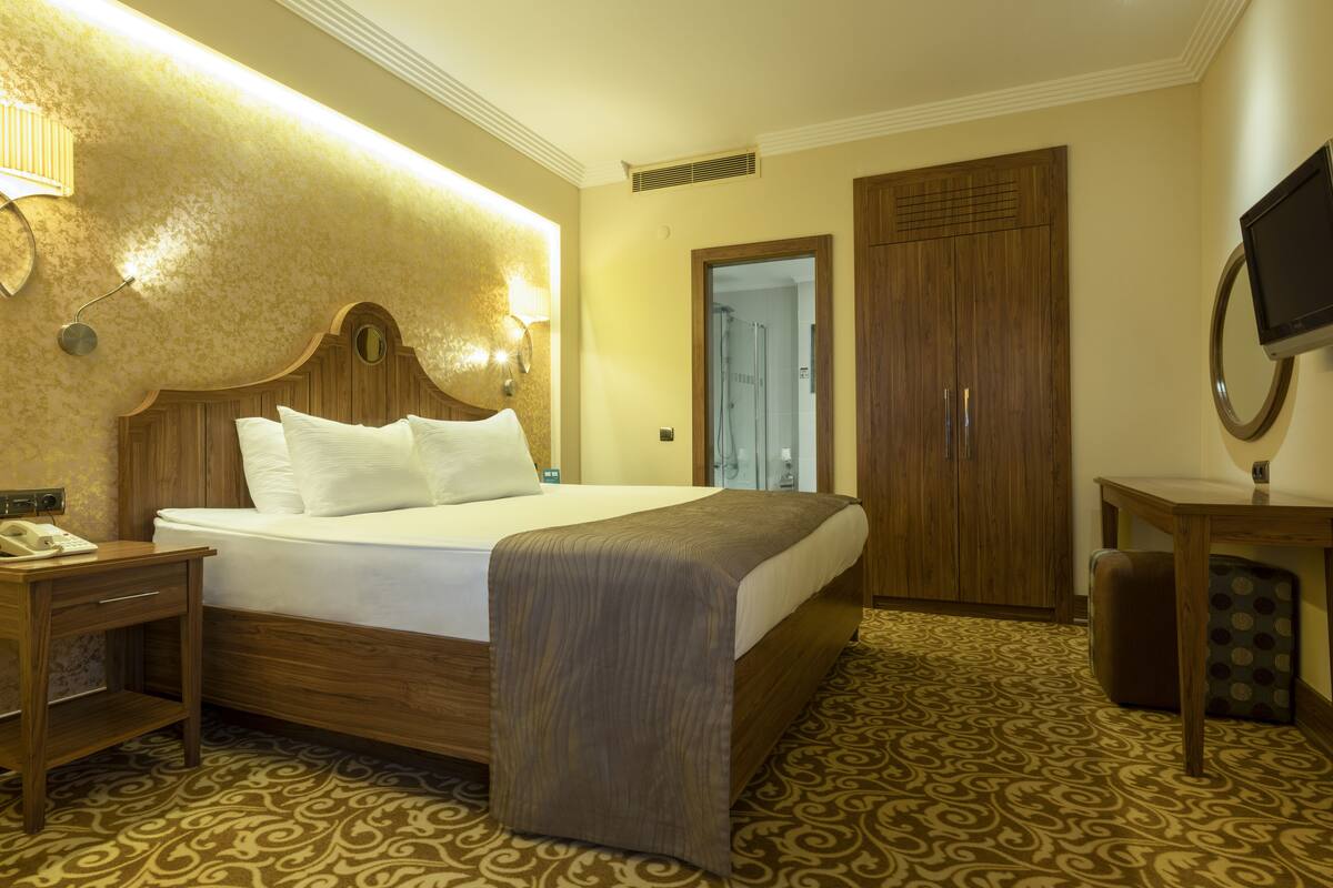 A hotel room with a large bed, wooden headboard, bedside table with a telephone, a television on a stand, and a wardrobe.