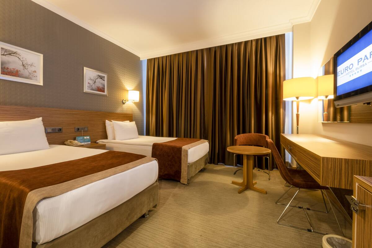 A hotel room with two beds, a desk, a chair, a television, and a wall-mounted lamp.