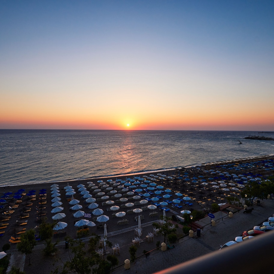 A beach with rows of umbrellas and lounge chairs, a sunset in the background, and a view of the sea.