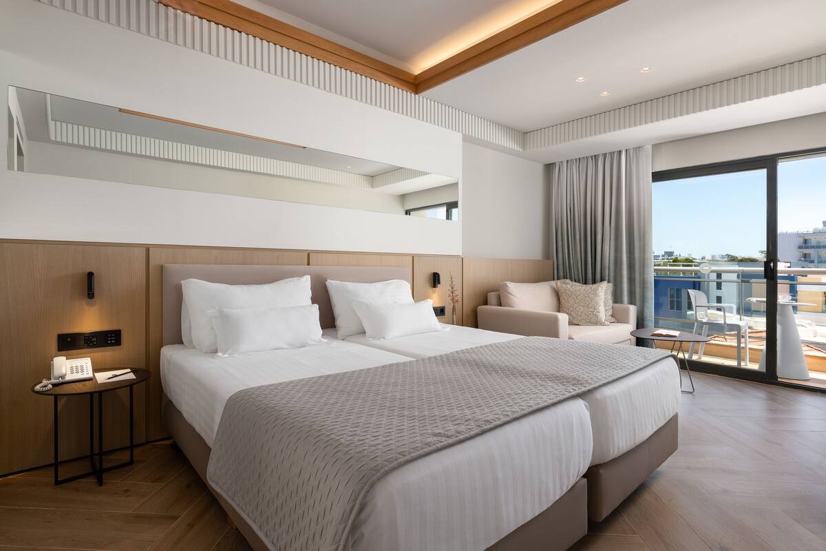 A modern hotel room with a large bed, a small desk, a seating area, and a balcony with a city view.