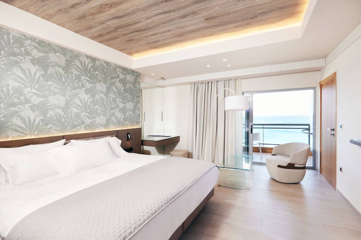 A modern hotel room with a large bed, a desk, a chair, and a balcony with a view of the sea.