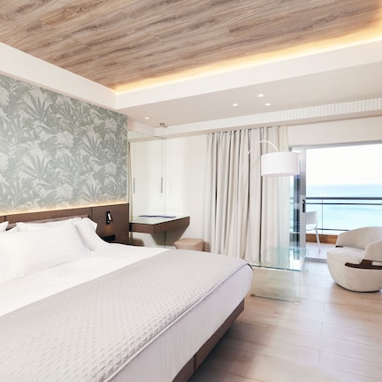 A modern hotel room with a large bed, a desk, a chair, and a balcony with a view of the sea.