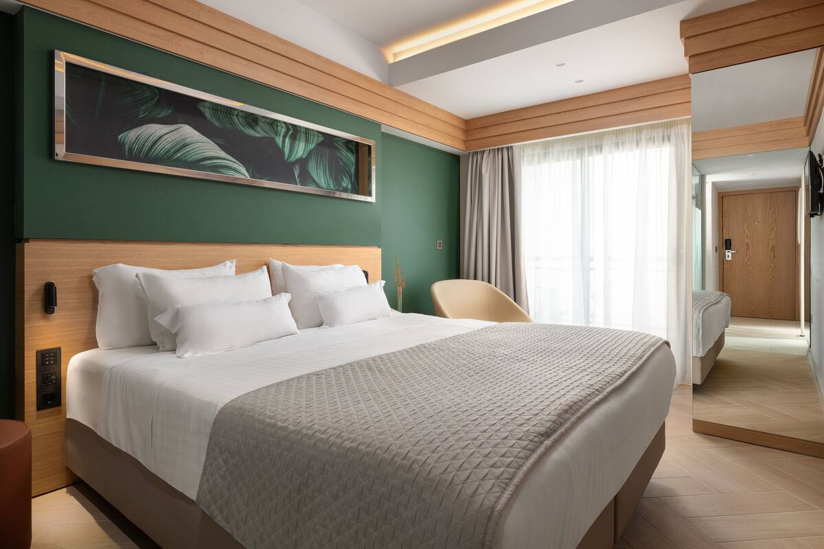 A modern hotel room with a large bed, a wooden headboard, a green accent wall, and a large window with curtains.