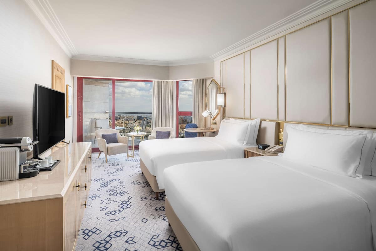 A hotel room with two beds, a desk with a TV, a small table with chairs, and a view of the city.