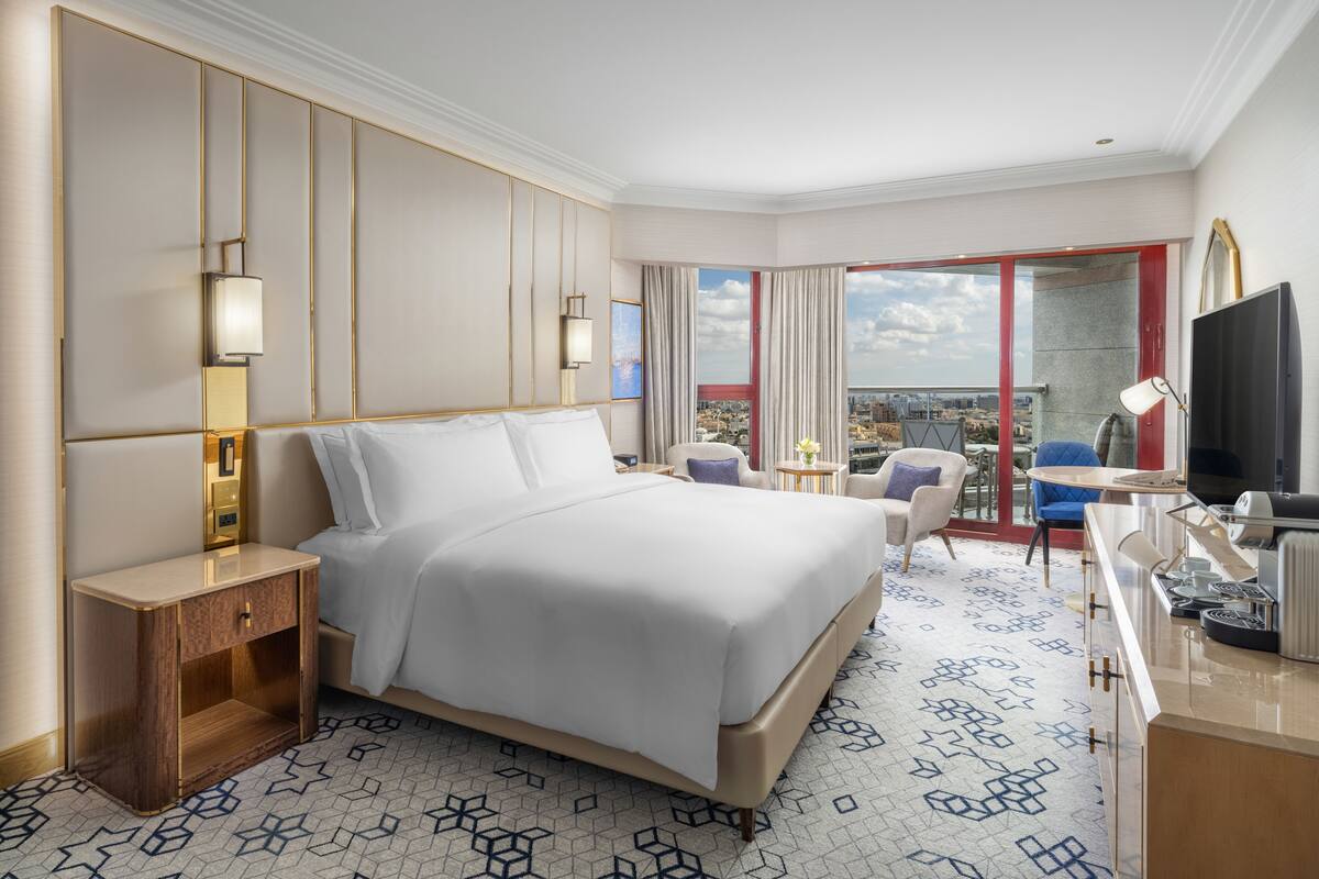 A hotel room with a large bed, a desk with a flat-screen TV, a chair, a small table, and a view of the city.