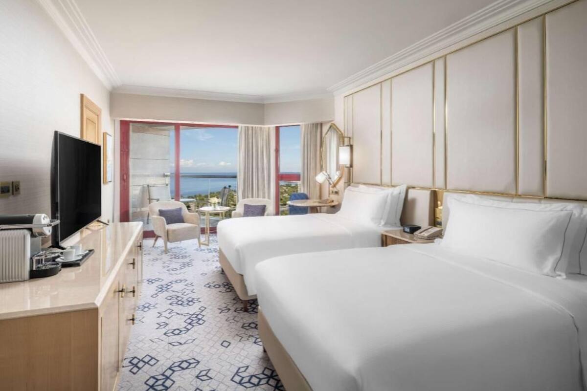 A hotel room with two beds, a TV, a desk with a lamp, and a view of the sea.