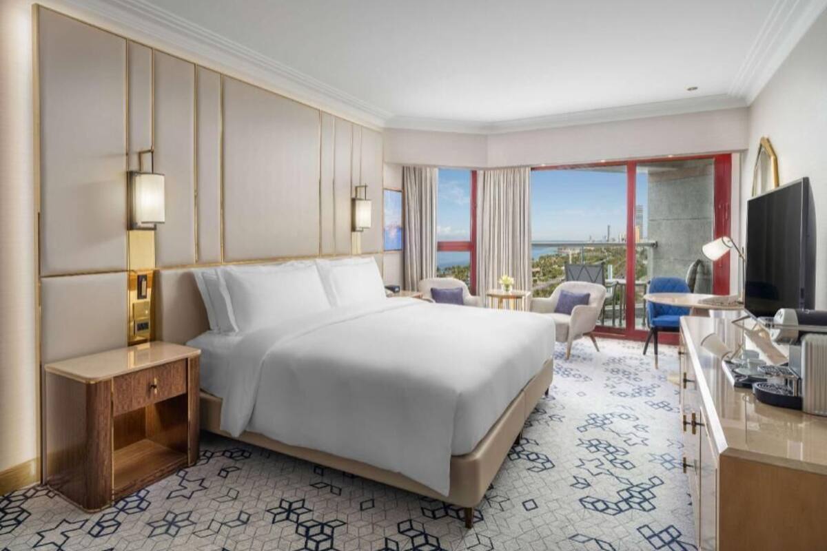 A hotel room with a large bed, bedside tables, a desk with a chair, a television, and a view of the city.