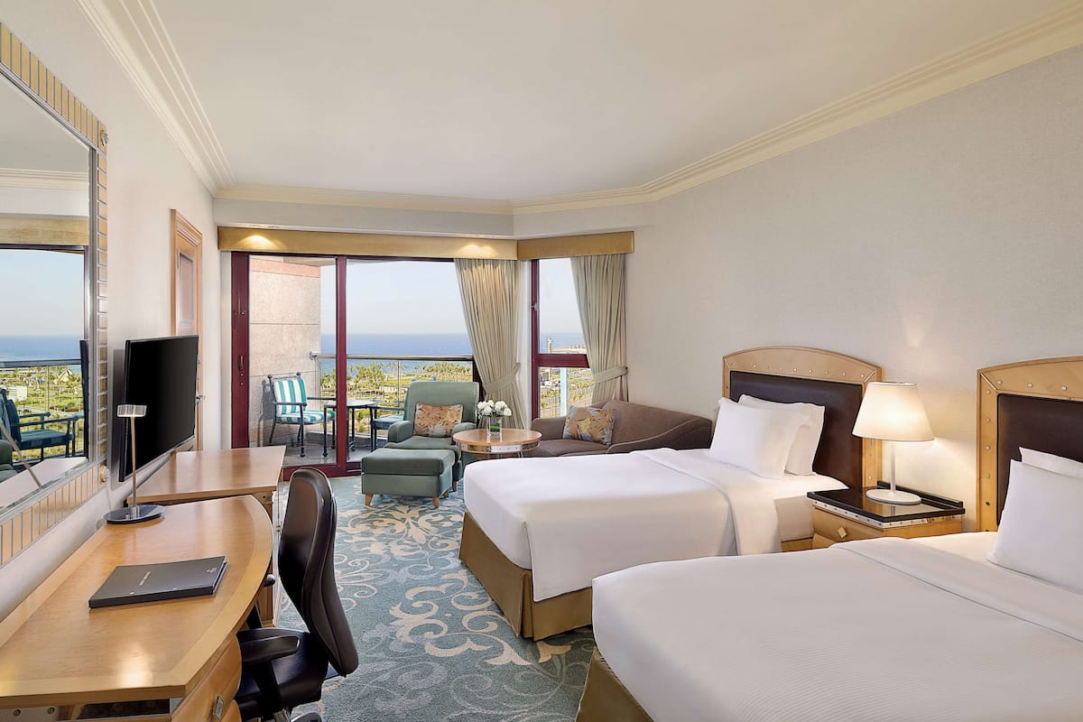 A hotel room with two beds, a desk, a chair, a TV, and a balcony with a view of the ocean.