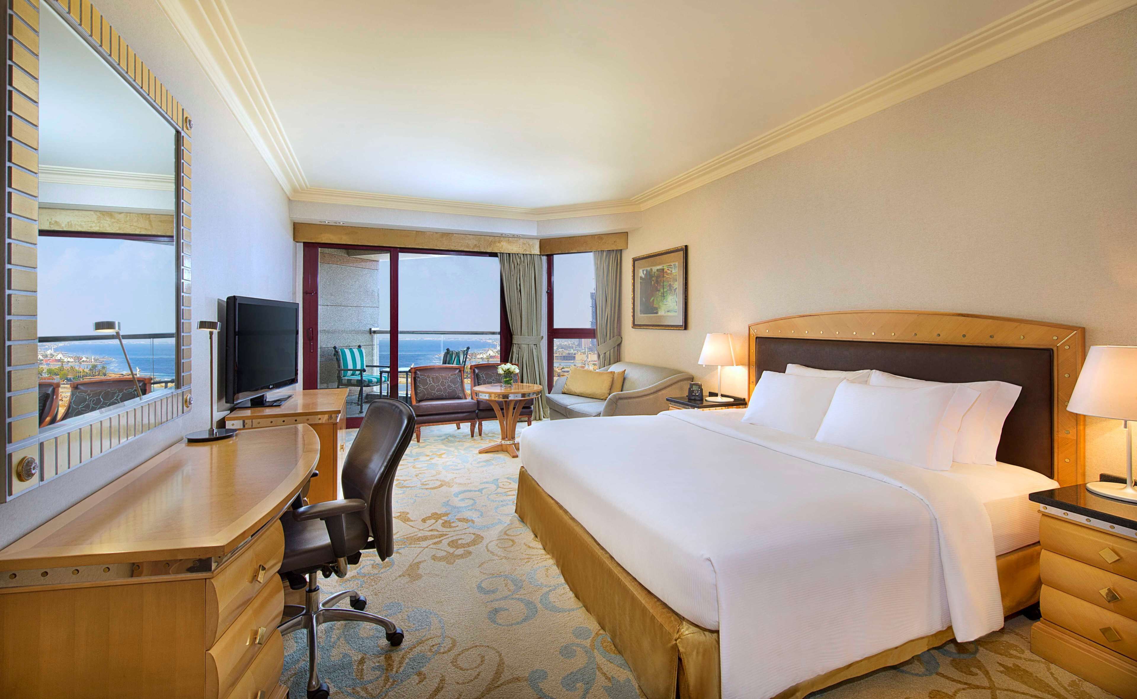 A hotel room with a large bed, a desk with a TV, a chair, a table with a vase, and a view of the ocean.
