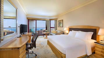 A hotel room with a large bed, a desk with a TV, a chair, a table with a vase, and a view of the ocean.