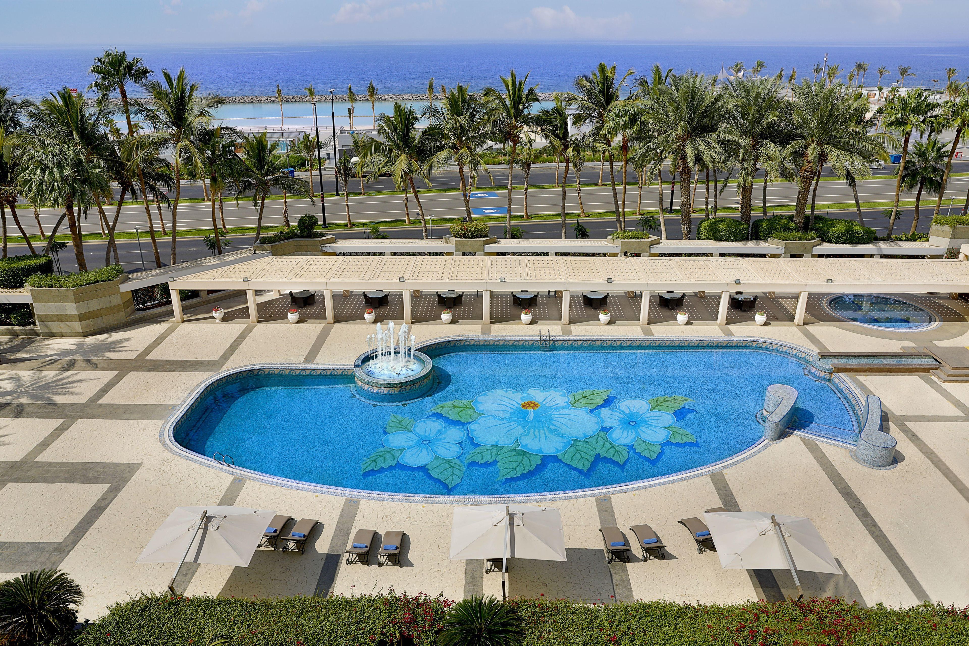A swimming pool with a fountain, surrounded by lounge chairs and umbrellas, with a view of the ocean and palm trees.