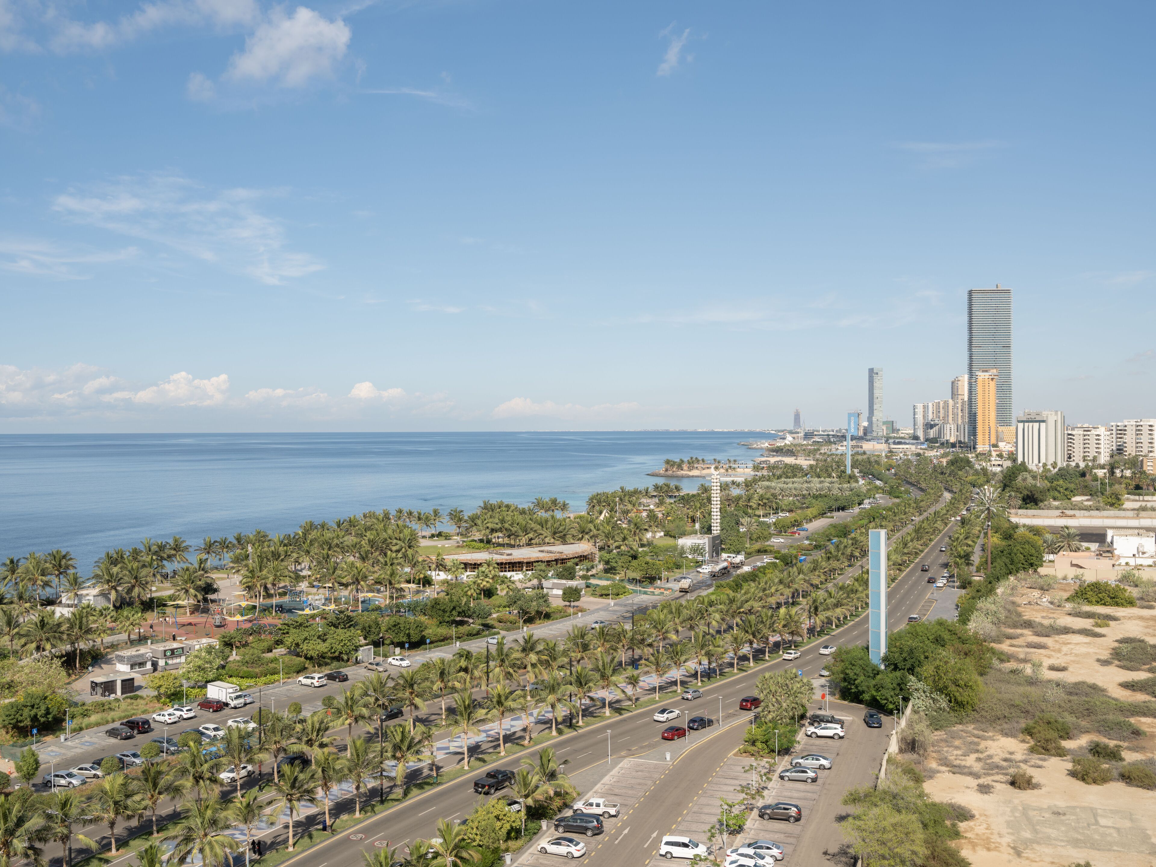 Un paisaje urbano costero con una torre alta destacada, una avenida concurrida bordeada de palmeras y una vista despejada del océano.