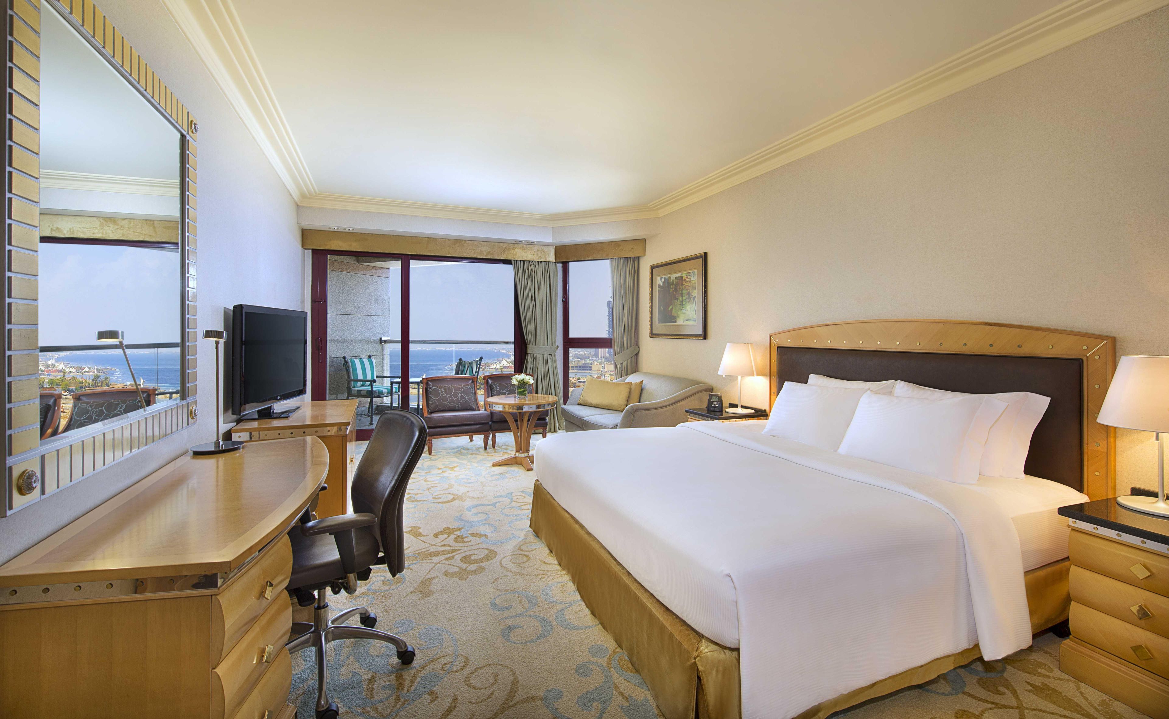 Ein Hotelzimmer mit einem großen Bett, einem Schreibtisch mit Computer, einem Sessel, einem Fernseher und Blick aufs Meer.