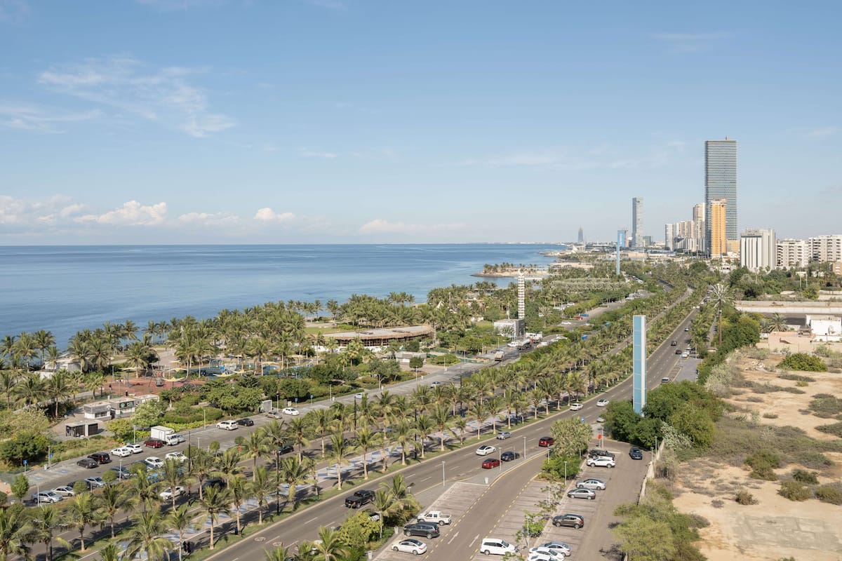 A coastal cityscape with a prominent tall tower, a busy road lined with palm trees, and a clear view of the ocean.