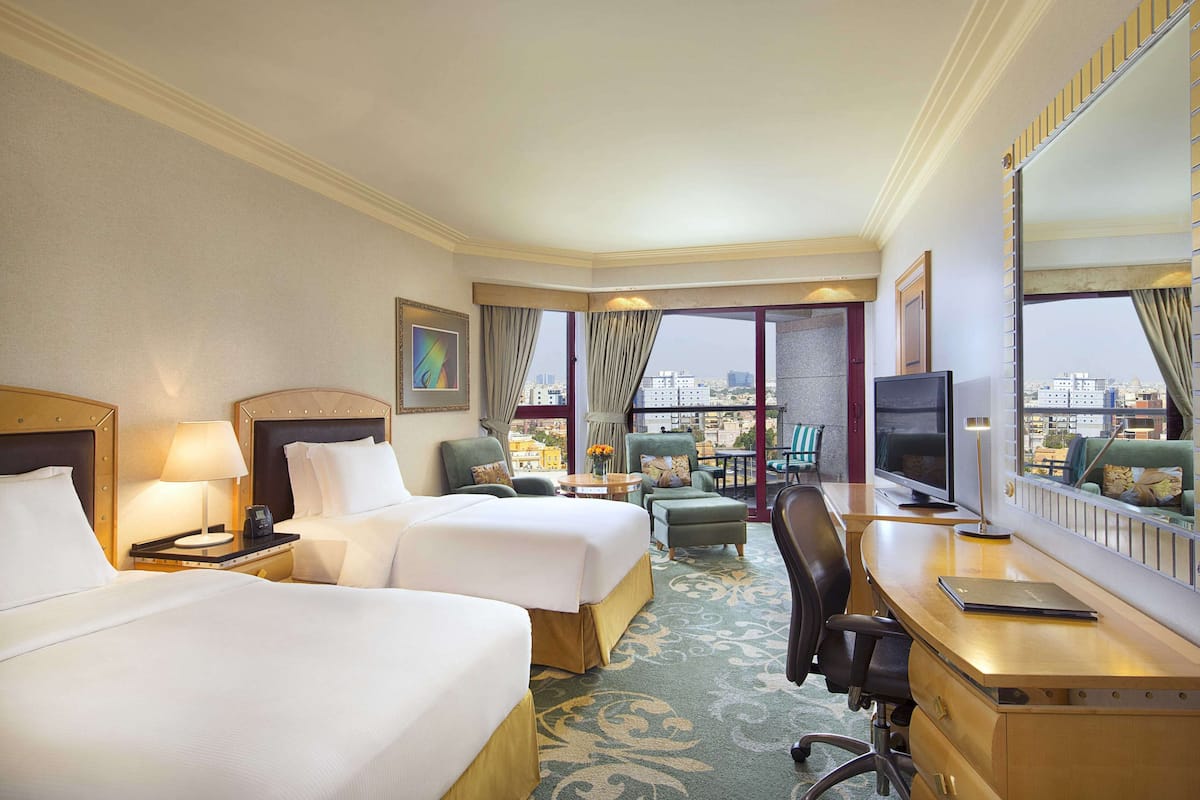 A hotel room with two beds, a desk, a chair, a TV, and a large window with a city view.