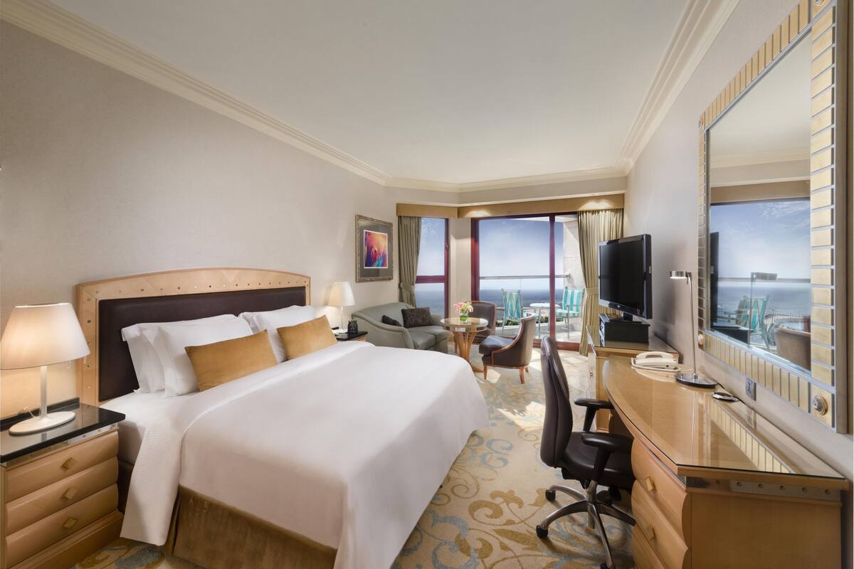 A hotel room with a large bed, a desk with a computer, a TV, and a view of the ocean.