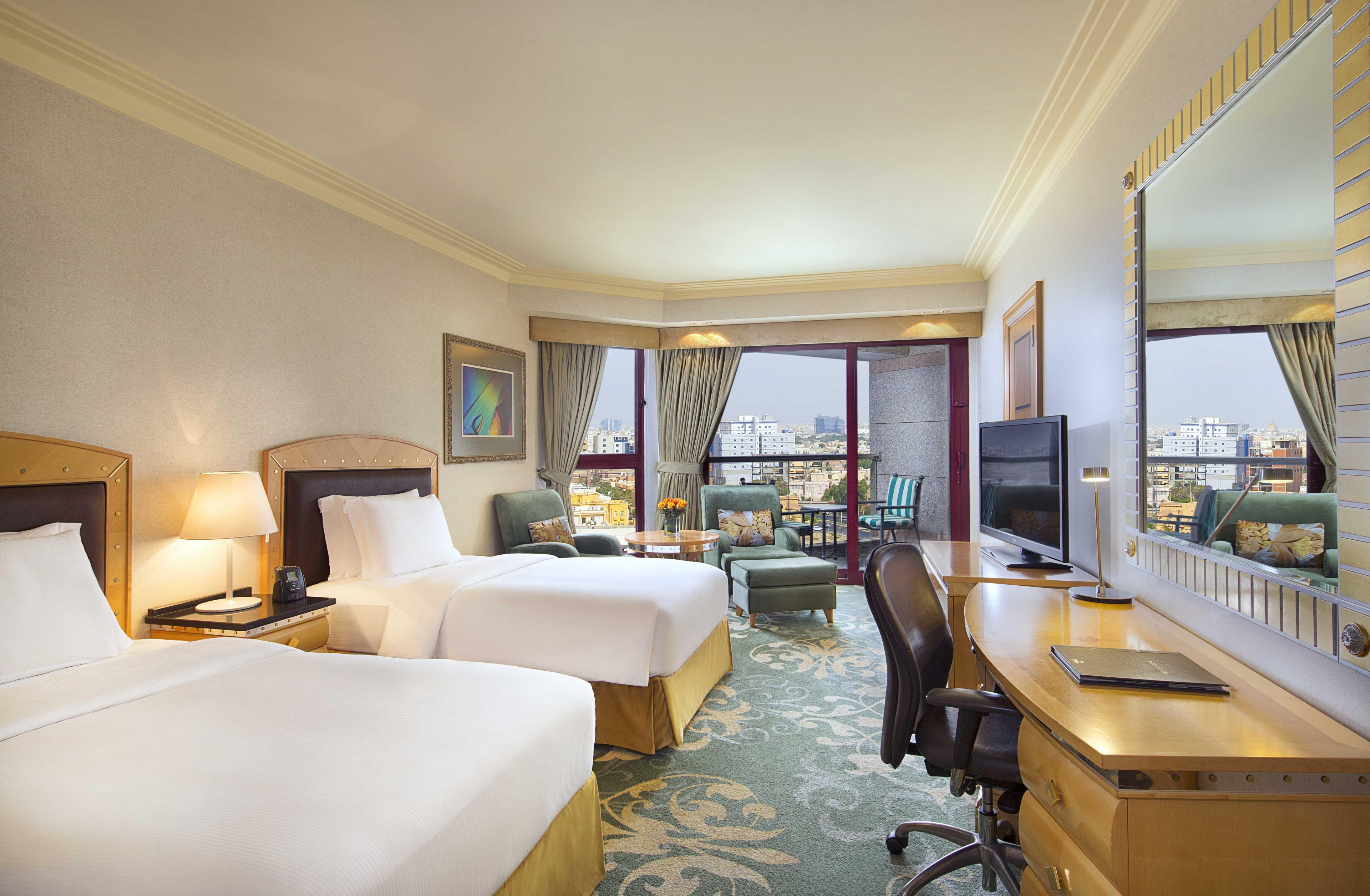 A hotel room with two beds, a desk, a television, and a view of the city.