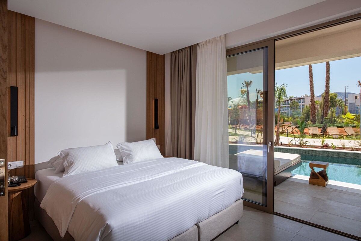 A modern hotel room with a large bed, a bedside table, a TV, and a view of a swimming pool and palm trees.