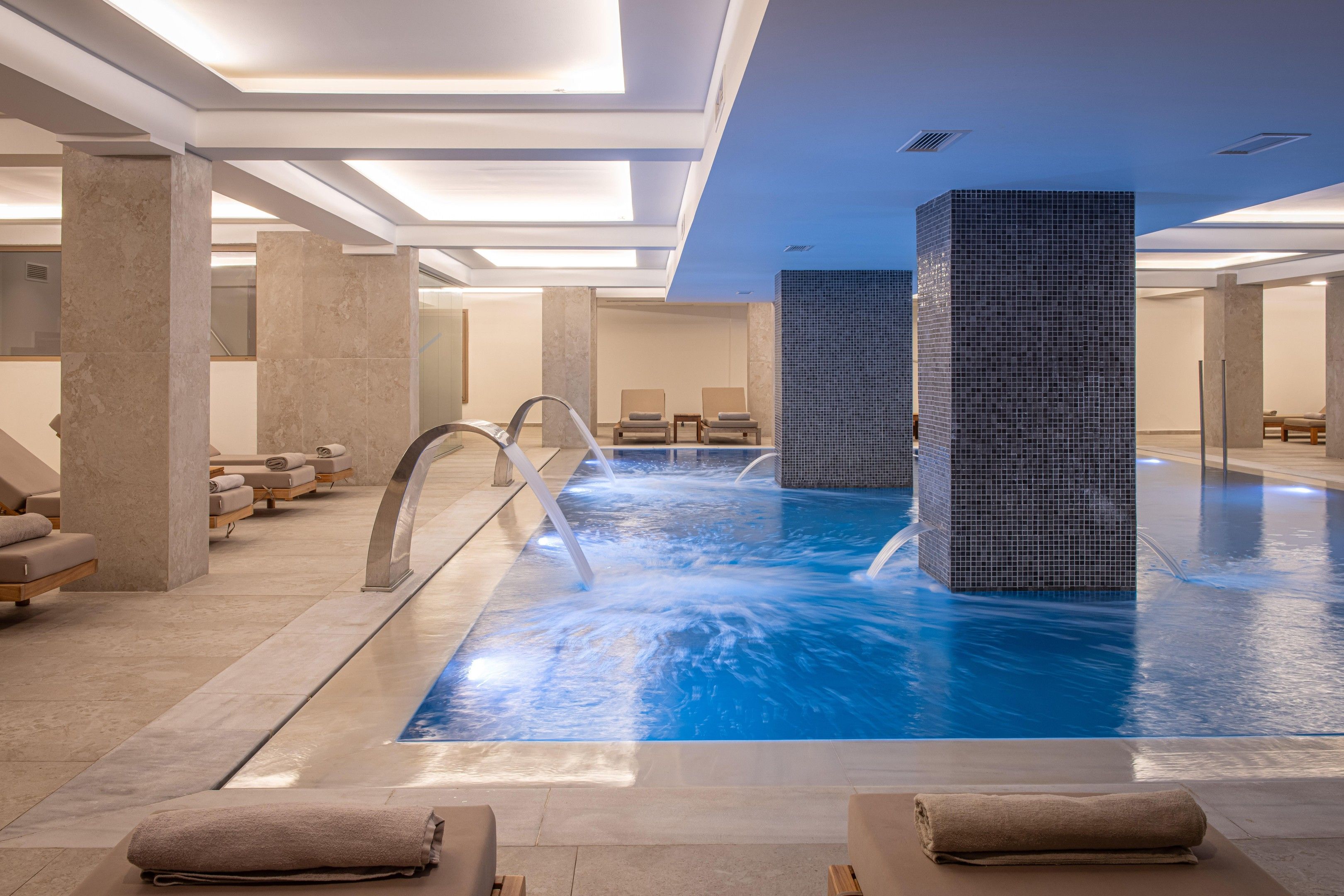 Piscina interior moderna con agua cristalina, rodeada de sillones de descanso y un techo alto con iluminación empotrada.