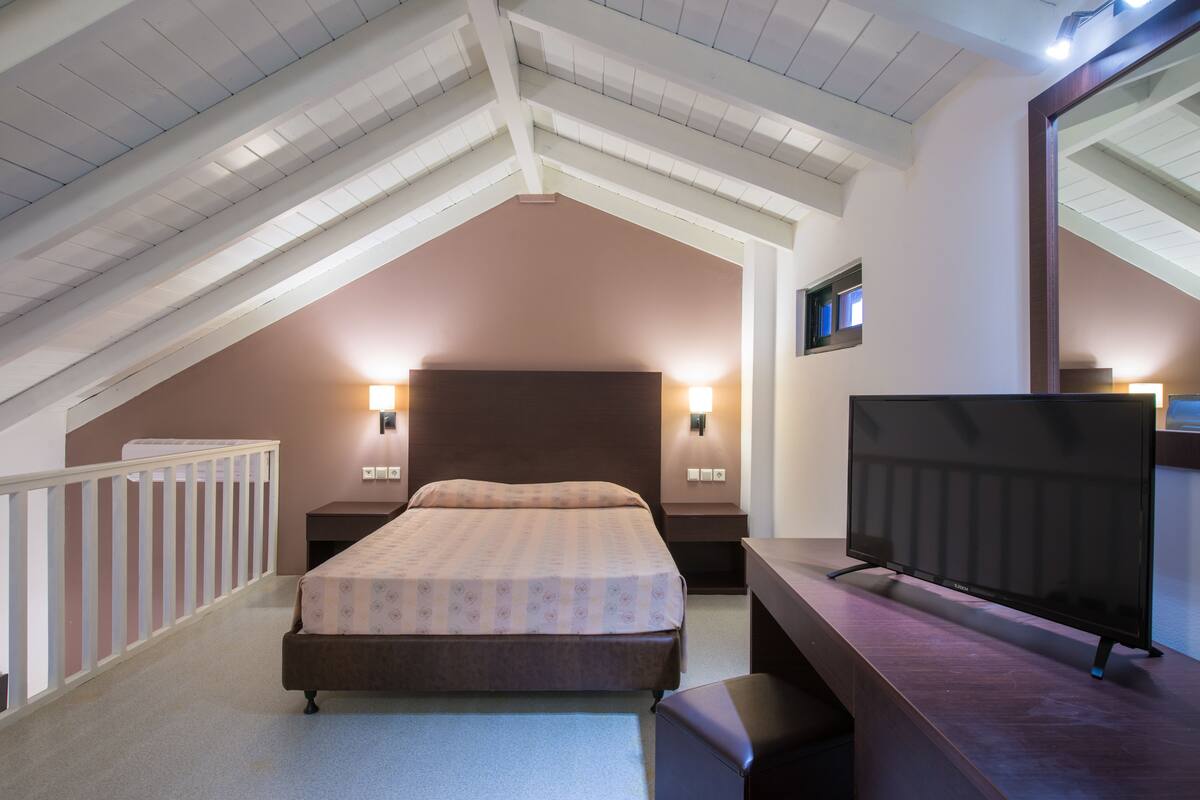 A bedroom with a sloped ceiling, a bed, bedside lamps, a TV, and a mirror.