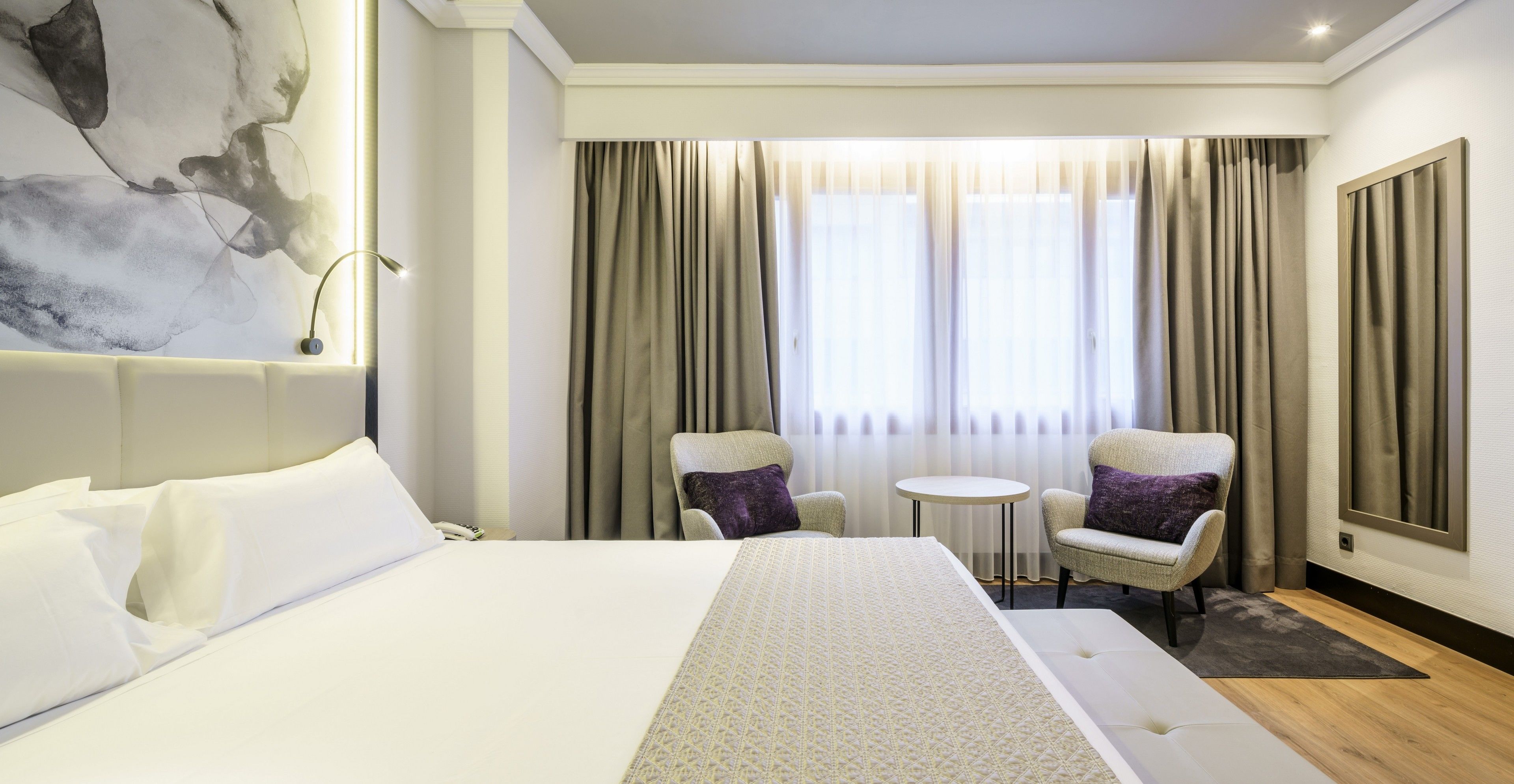 A hotel room with a large bed, two armchairs, a small round table, and a large window with curtains.