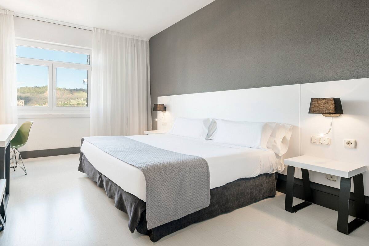 A modern hotel room with a large bed, a desk, and a chair. There is a window with curtains, a lamp, and a view of the outdoors.