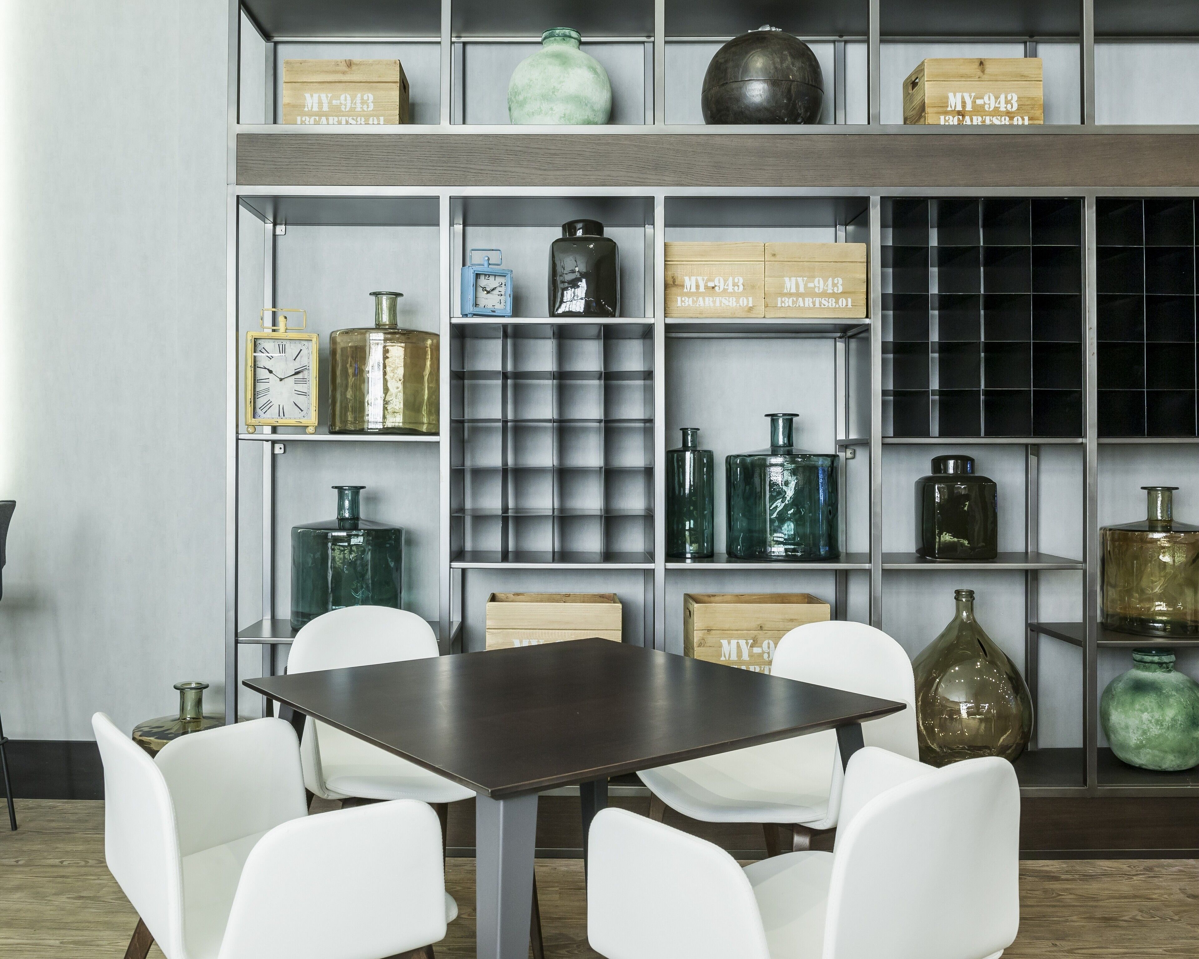 A modern interior with a dark wooden table and white chairs, surrounded by shelves with various glass bottles and wooden boxes labeled "MY-943".