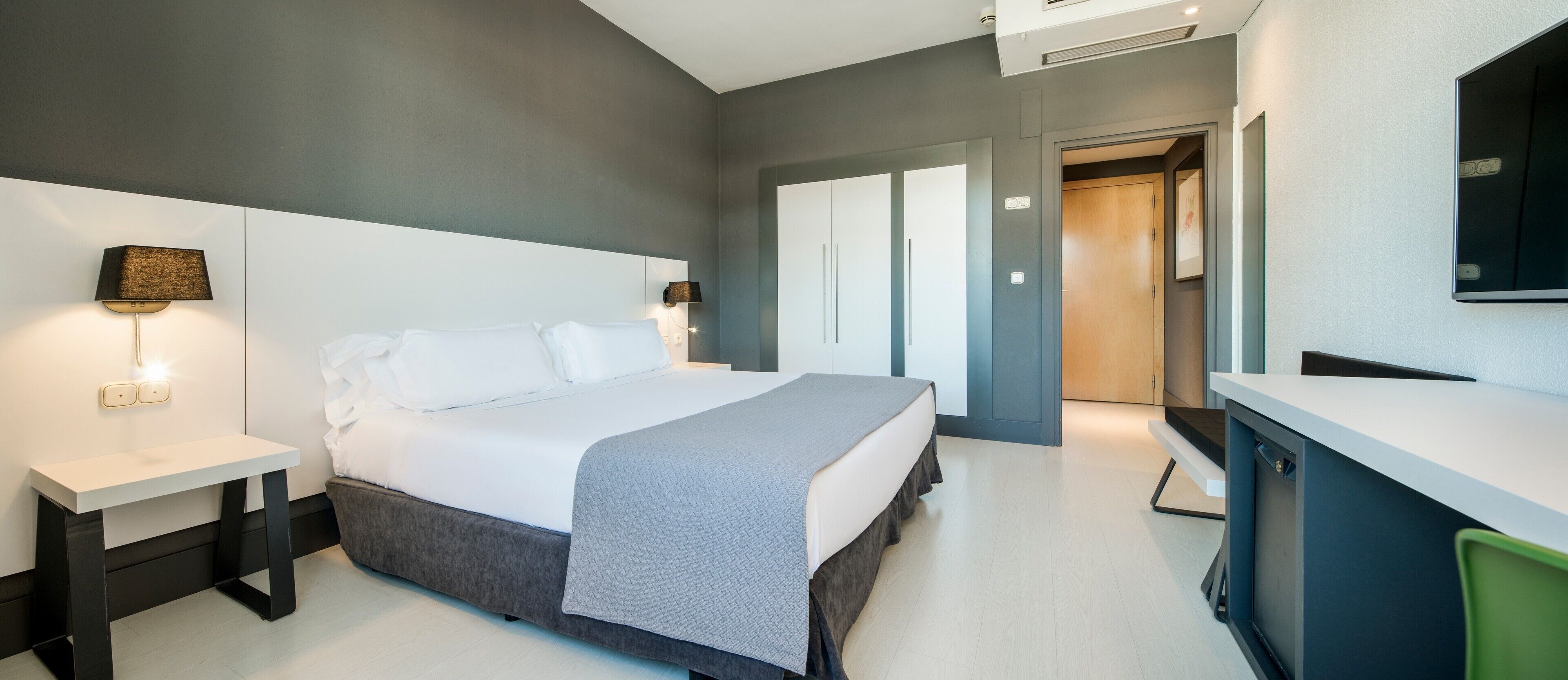 Una habitación de hotel moderna con una cama grande, mesitas de noche, un escritorio y un espejo.