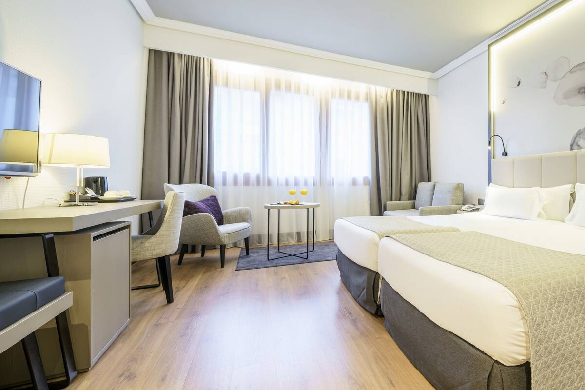 A hotel room with a bed, two chairs, a desk, a small table, and a large abstract painting on the wall.