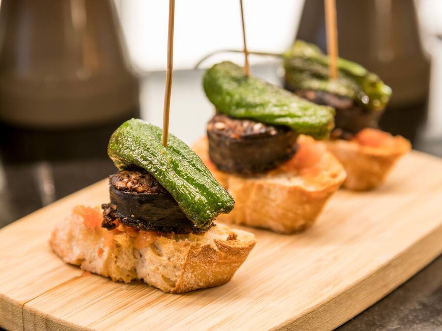 Una tabla de madera con alimentos, entre ellos una rebanada de pan cubierta con un vegetal a la parrilla y un trozo pequeño de carne.