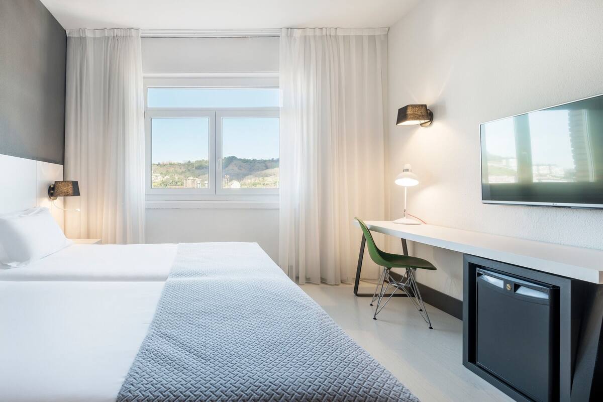 A hotel room with two beds, a desk, a TV, and a window with a view.