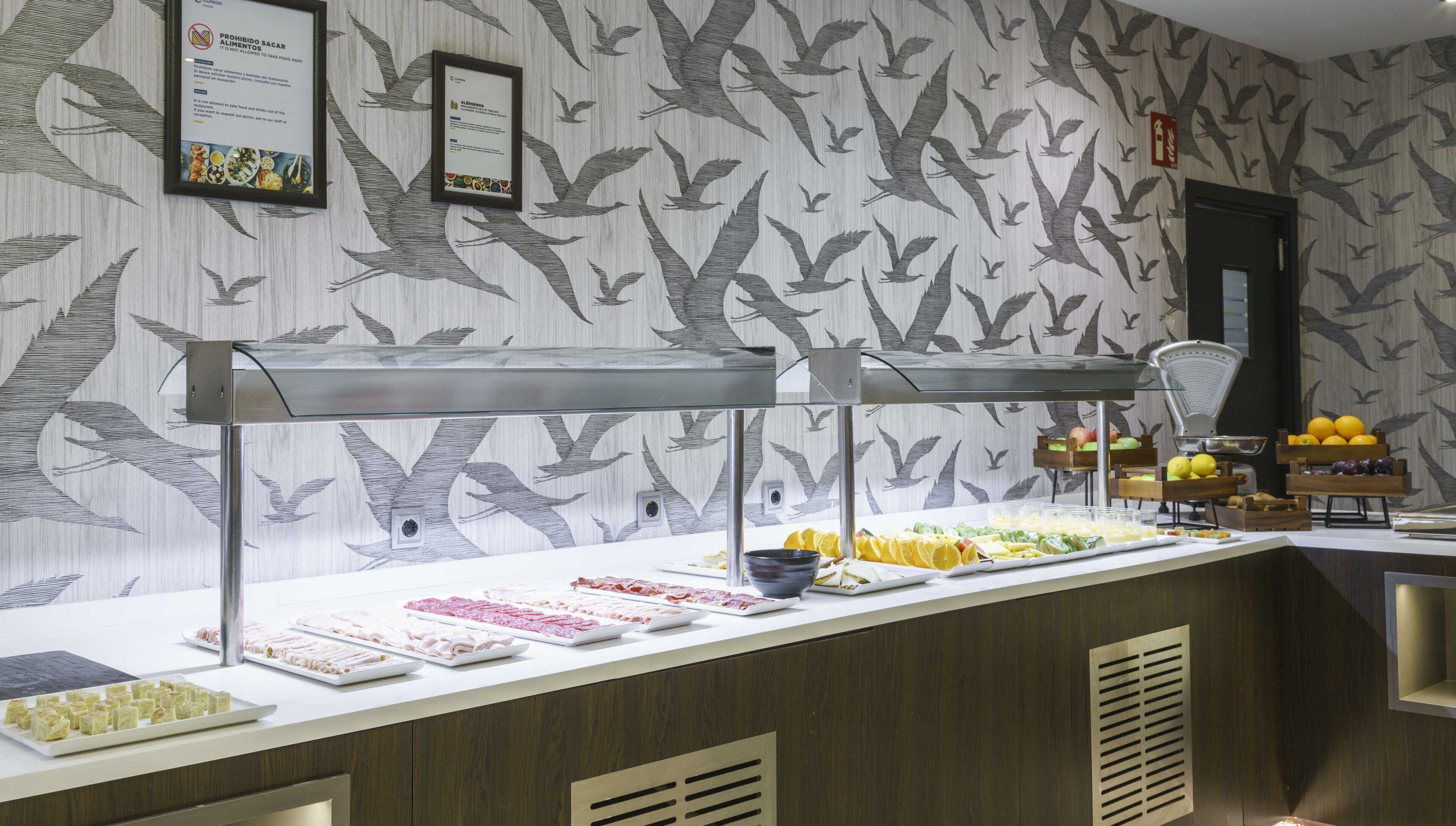 Un espace buffet proposant une variété de plats, notamment des fruits et des viandes, le tout disposé devant un mur décoré d’un motif représentant un arbre et des oiseaux.