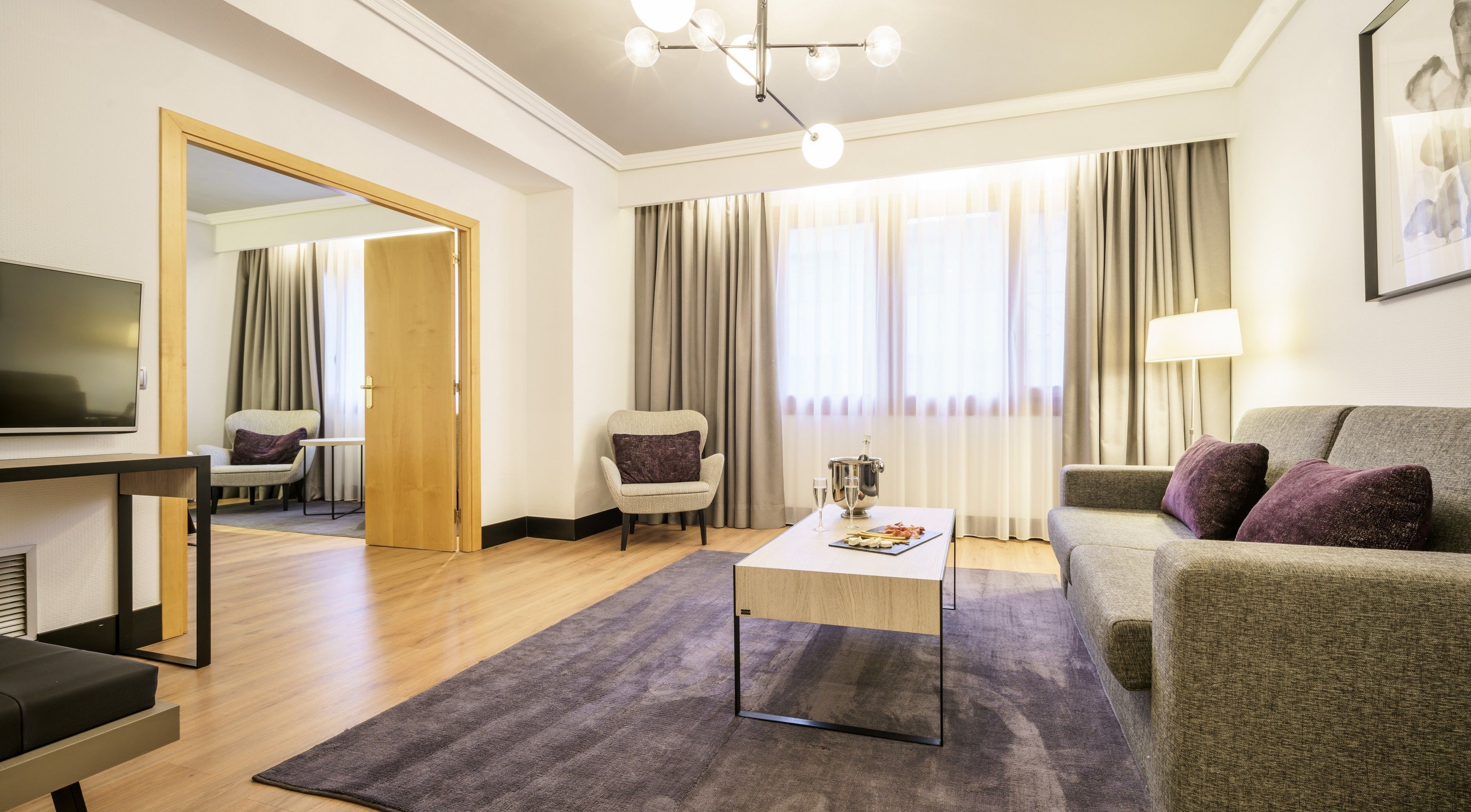 A modern hotel room with a flat-screen TV, a sofa, a chair, a coffee table, and a large window with curtains.