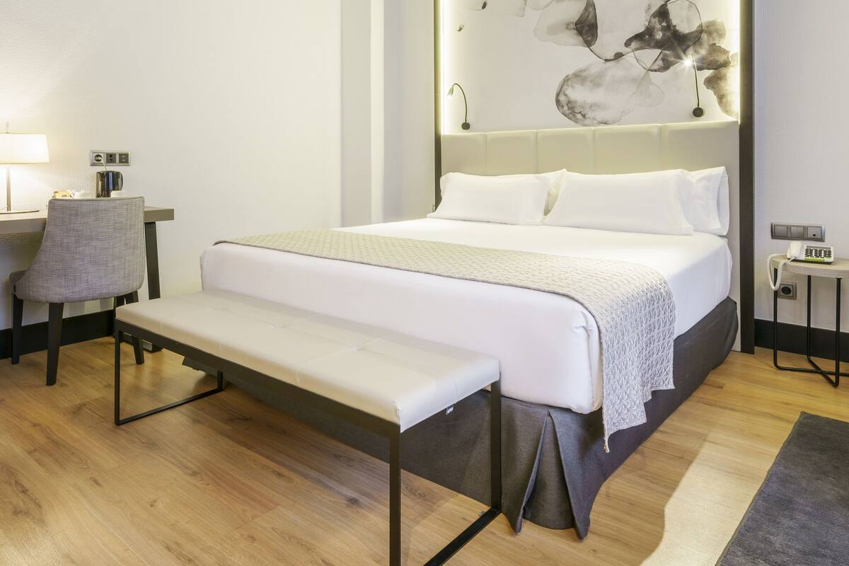 A hotel room with a bed, a desk with a chair, a television, and a bedside table with a lamp.