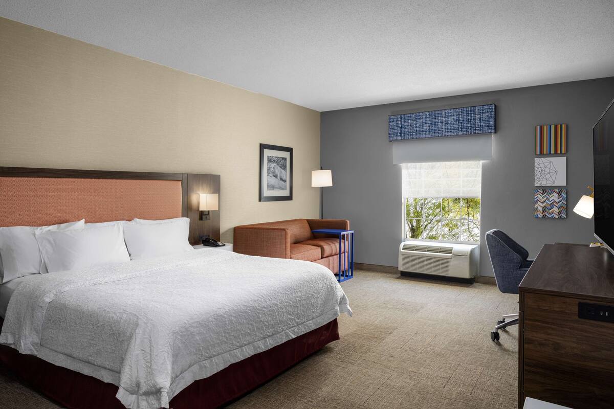 A hotel room with a large bed, a desk, a chair, a window with a view, and a wall with artwork.