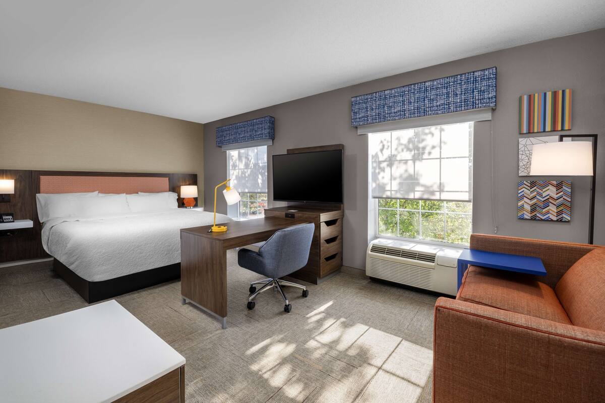 A hotel room with a bed, desk, chair, TV, and large windows.