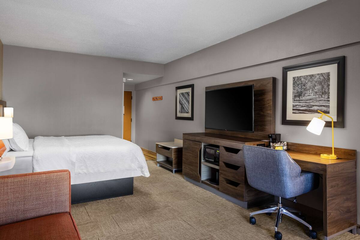 A hotel room with a bed, a desk, a chair, a TV, and artwork on the wall.