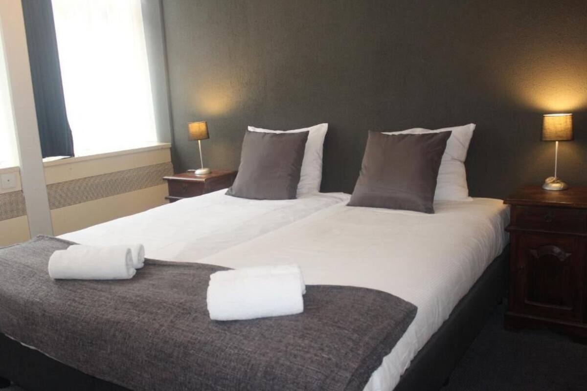 A neatly made bed with white and gray linens, two bedside lamps, and a window with curtains.