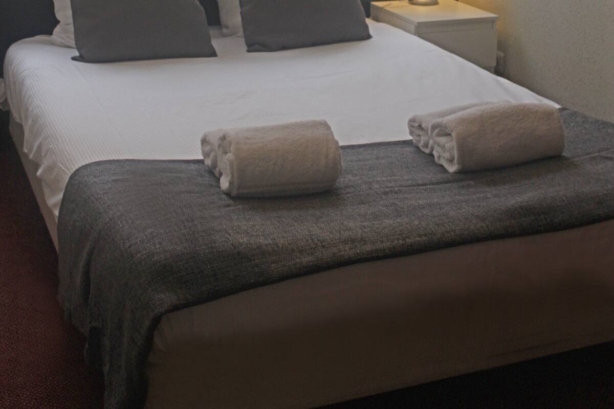A neatly made bed with pillows and a blanket, a bedside lamp, and a window with curtains.