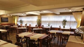 Een restaurant met meerdere tafels en stoelen, grote ramen met gordijnen en een aan de muur bevestigde televisie.