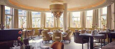 Un restaurant spacieux avec des tables et des chaises rondes, de grandes fenêtres et un lustre central.