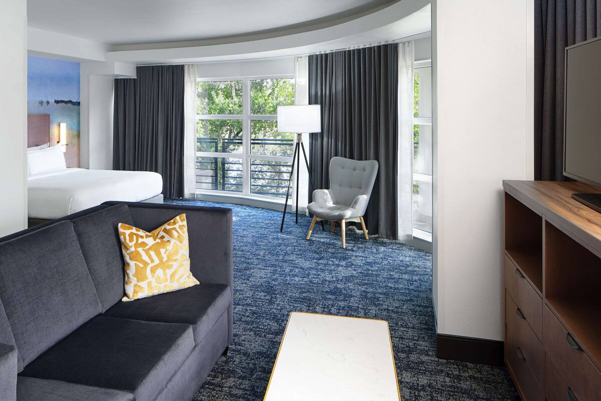 A hotel room with a grey sofa, a white bed, a small armchair, a TV stand, and a large window with curtains.