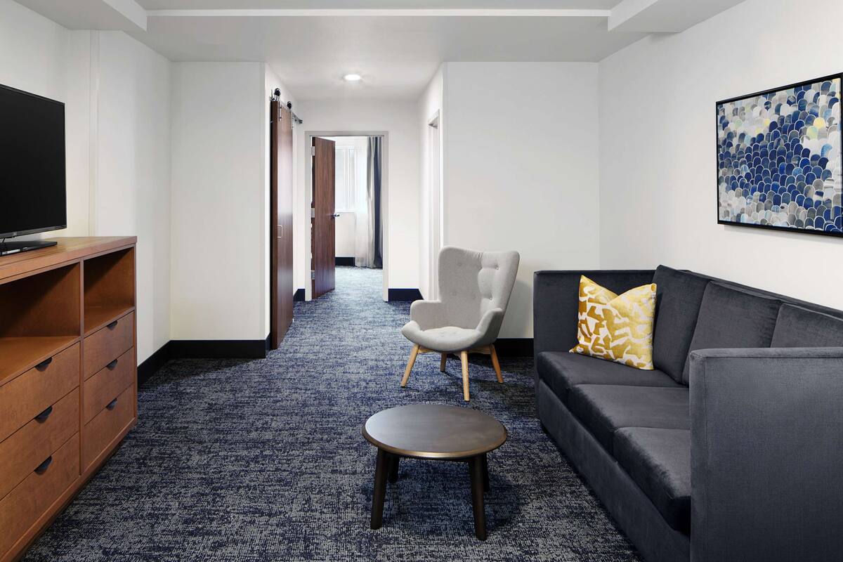 A modern hotel room with a flat-screen TV, a wooden cabinet, a grey sofa, a small round table, and a hallway leading to a bathroom.